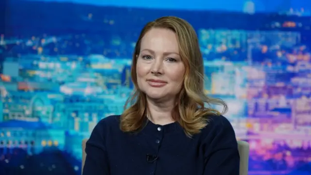 Chauntae Davies, with blonde shoulder-length hair, wearing a navy jumper, looks straight ahead. A blue and purple cityscape sits in the background.