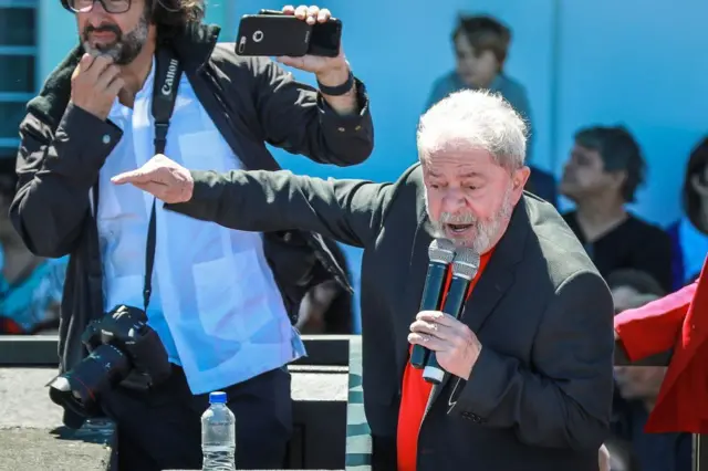 Lula da Silva vistiendo una chaqueta oscura y una camisa roja pronuncia un discurso desde una tarima mientras carga dos micrófonos en la mano izquierda. Detrás de él, a la izquierda, hay un hombre con una cámara fotográfica que le cuelga del cuello y que parece estar grabando a Lula con un celular.