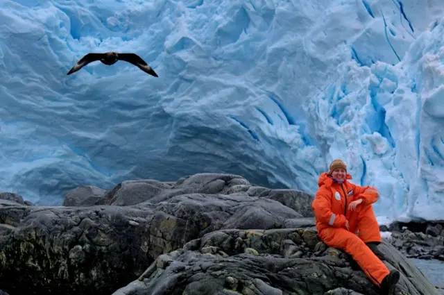 Анжеліка Ганчук в помаранчевому комбінезоні сидить на скелястому березі на фоні снігів