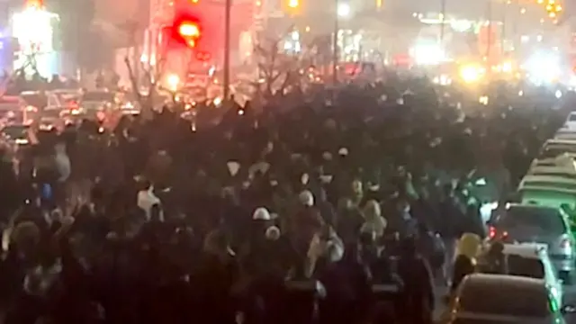 Đám đông nhìn từ phía sau đang tuần hành dọc theo một con phố ở Tehran vào đêm ngày 8/1/2026. Bên phải người biểu tình là một hàng xe hơi, bên trái là nhiều làn xe cộ khác, phía xa là những ánh đèn sáng rực.