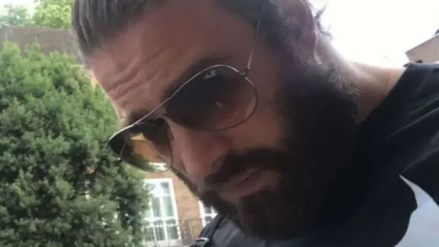 A photo of Chris Harkins from his Tinder profile. He is outside with trees and houses in the background. He has his dark hair slicked back and a full beard. He is wearing large tinted sunglasses with a thin metal rim. He is looking down towards the ground. 