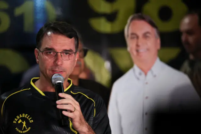 Flavio Bolsonaro discursa ao microfone, com imagem do seu pai atrás, em papelão