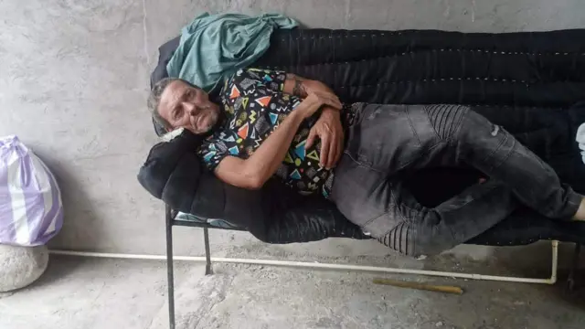 Lázaro Romero León mira a cámara tumbado en una silla alargada, con jeans gris oscuros y una camiseta estampada, en un patio de cemento en Tapachula, Chiapas, México.