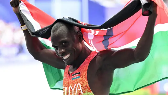 Emmanuel Wanyoni, wearing a brightly coloured Kenya running vest, smiles broadly as he raises a Kenyan flag behind him in celebration at the Paris 2024 Olympics