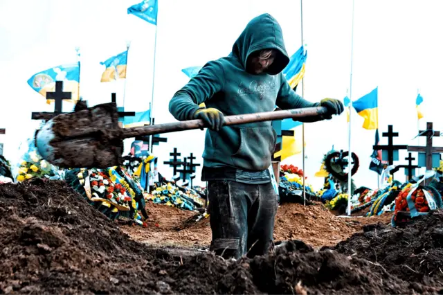 2024年3月16日,掘墓工人在敖德萨的一座墓园挖掘坟墓。他们身后约有十面乌克兰国旗飘扬在白色旗杆上。前景中的一名掘墓工人身穿青绿色连帽上衣,戴着黄色手套