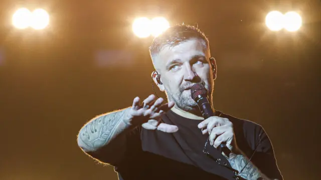 Мужчина в черной футболке, с микрофоном и татуировками на руках