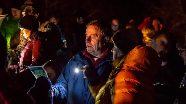 Primer plano de una multitud cantando y sosteniendo una partitura a la luz de una linterna. La luz les ilumina el rostro. Llevan abrigos de invierno coloridos, como naranja, azul y verde.