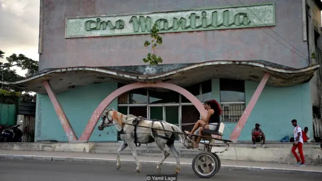 Casal em charrete passa em frente a cinema com aspecto envelhecido em Havana