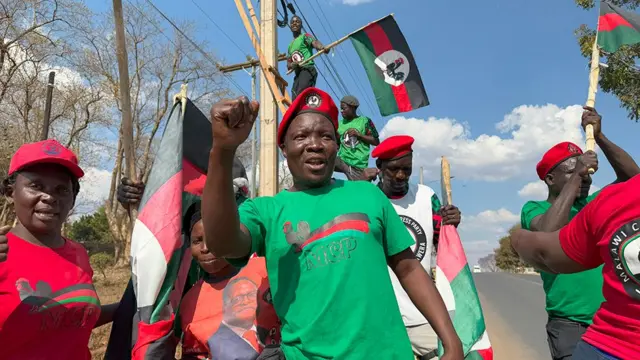 Des partisans du MCP de Lazarus Chakwera sur le bord de la route. Le personnage principal, au centre, lève le poing et porte un t-shirt vert du parti et un béret rouge. D'autres brandissent des drapeaux du parti.
