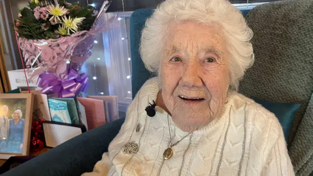 Elsie Wilkins sonríe a la cámara, sentada en un sofá. Lleva un jersey de punto trenzado color crema con un broche brillante y un colgante dorado. Detrás de ella, sobre una mesa, hay un ramo de flores y tarjetas.