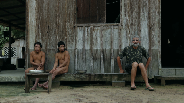 Dois homens indígenas ao lado de um homem branco em frente a uma casa