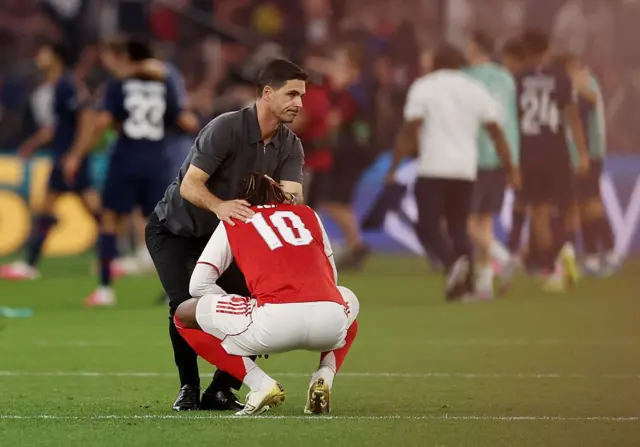 Umutoza wa Arsenal Mikel Arteta aciye bugufi arimo guhoza umukinnyi we Eberechi Eze, usutamye yubitse umutwe ateye umugongo ‘camera’, nyuma yuko Arsenal itsinzwe kuri penaliti na Paris Saint-Germain, ku mukino wa nyuma wa UEFA Champions League, ku kibuga Puskas Arena, ku wa gatandatu, ku itariki ya 30 Gicurasi (5) mu 2026, i Budapest, mu murwa mukuru wa Hongrie.