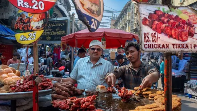 Mumbai'de tavuk tikka reklam tabelasının yanında, pişmiş etlerin bulunduğu büyük metal standların yanında duran iki adam, 2022