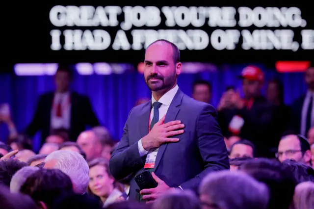 Eduardo Bolsonaro, um homem branco de terno, barba e bigode curto, ficando calvo, com a mão no peito em meio a uma multidão