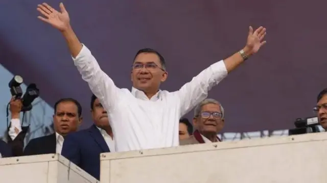 طارق الرحمان په سپينو کالیو کې پر دريځ ولاړ دی، لاسونه یې پورته کړي دي او شاوخوا یې نور چارواکی هم برېښي. 