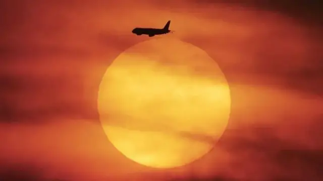 کره کامل خورشید یا ماه به رنگ زرد در هوایی قرمز رنگ، پرهیب هواپیمایی در حال پرواز که در مقابل کره خیلی کوچک است دیده می‌شود