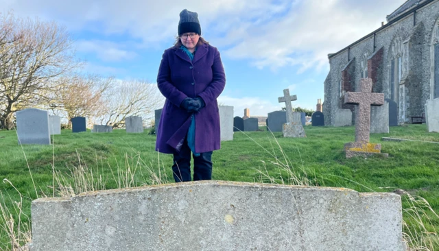 Una mujer con un abrigo morado de botonadura sencilla, un gorro de lana azul marino y pantalones negros observa una lápida, de pie frente a la cámara. Sus manos enguantadas están entrelazadas. Detrás de ella hay otras lápidas y el lateral de una iglesia.   