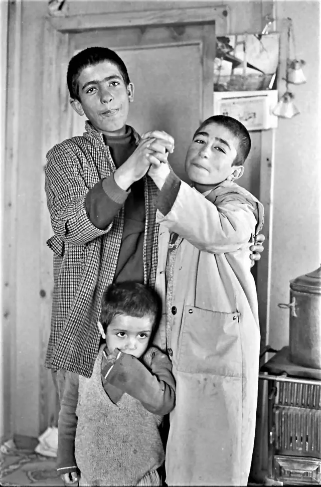 Üç çocuk birlikte görülüyor. Çocuklardan yaşları daha büyük olan ikisi, dans etme pozu veriyor. Daha küçük çocuk ise ortalarında duruyor. Arkalarında da üstünde kazan olan bir soba var. 