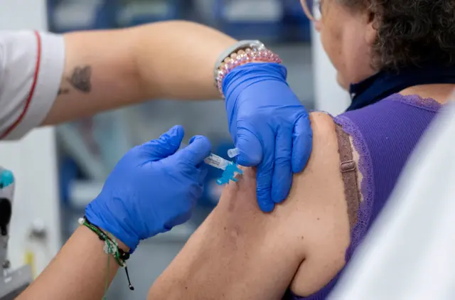 کارمند بهداشتی با دستکش آبی در حال تزریق واکسن به بازوی یک زن است.