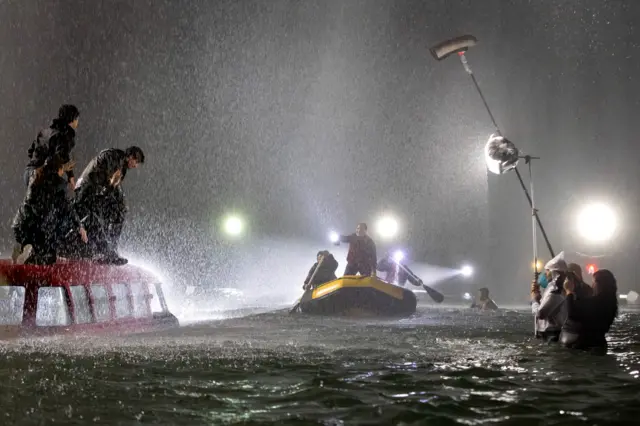  pessoas se equilibram sobre um carro parcialmente submerso enquanto um bote se aproxima, e, ao lado, a equipe opera luz e som dentro da água.