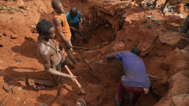 Group of men digging rubble looking for valuables, one no wear cloth while one wear blue cloth. Dem bend down and anoda wear brown singlet.