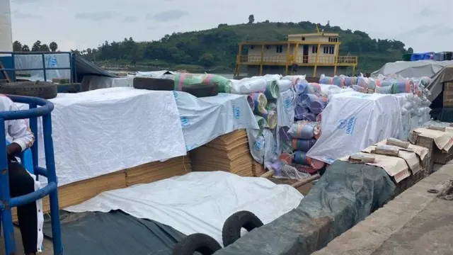 Ibicuruzwa biri mu cyambu cya Rubavu, birimo ibitwikirijwe shitingi, hakurya haboneka umusozi urimo ibimera bitoshye. 