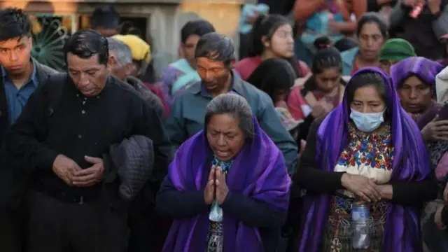 Na foto, um homem do lado esquerdo vestidojogar truco online gratispreto com as mãos unidas olha para baixo. Ao ldo dele, uma mulher com as mãosjogar truco online gratisposiçãojogar truco online gratisoração, vestida com um pano roxo sobre a roupa, ao lado, uma mulherjogar truco online gratismáscara, com um pano roxo igual sobre a cabeça. No fundo, várias pesssoas