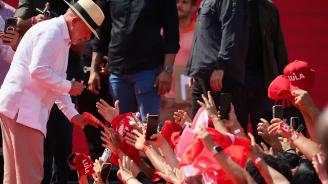 A imagem mostra Luiz Inácio Lula da Silva, de camisa e calça brancas e usando chapéu, inclinado para cumprimentar apoiadores em um evento. Na plateia, várias pessoas estendem as mãos e seguram celulares, bandeiras e bonés vermelhos com referências a Lula.
