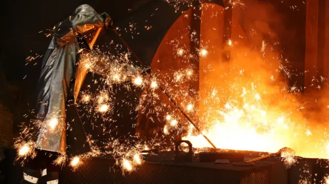 Sparks fly around a worker in a silver safety suit while he takes a sample of molten iron from a blast furnace in Duisburg, Germany