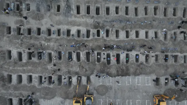 An aerial photo of at least five rows of rectangular holes in the ground, some marked and yet to be dug out, with three mechanical diggers visible.
