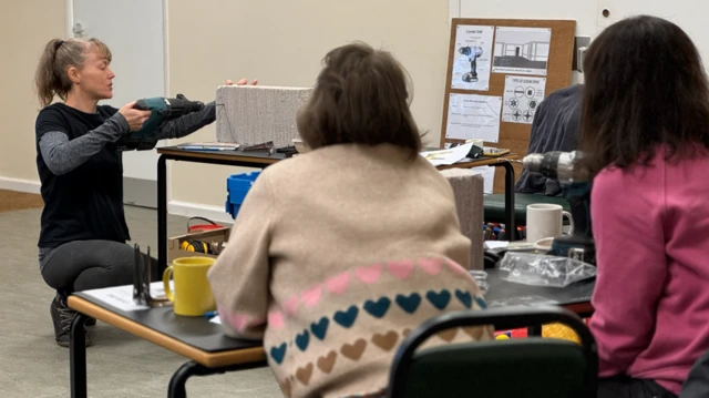 Sarah Bolger demuestra cómo usar un taladro en un bloque durante las sesiones de prueba. Está en cuclillas mientras hace la demostración, con el bloque sobre una mesa. Dos mujeres de espaldas a la cámara observan.