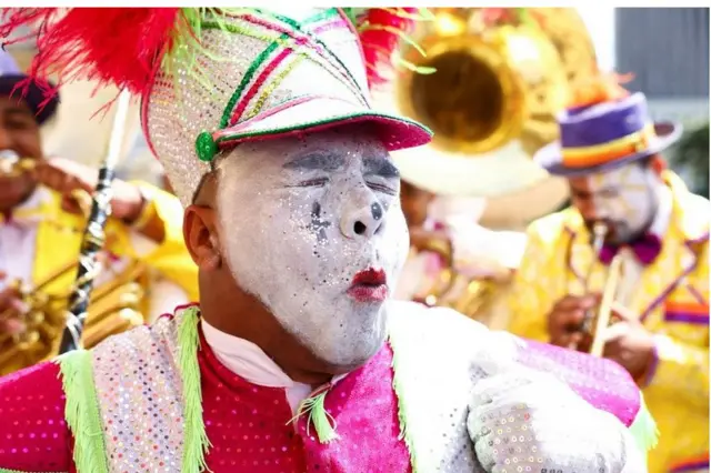 Wani mutum sanye da hula mai launuka daban-daban, ya shafa farin fenti a fuskarsa. Idanunsa a rufe, da yanayin fuska mai ban dariya, ya ranbada janbaki.