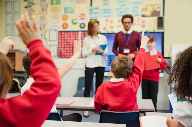 Uczeń szkoły podstawowej w brytyjskiej miejscowości Hexham prezentuje przed klasą. Na pierwszym planie inni uczniowie siedzący w ławkach i podnoszący ręce w górę. Uczniowie mają na sobie mundurki: czerwone bluzy i czarne spodnie. Na dalszym planie stoi dwójka nauczycieli: mężczyzna w bordowym swetrze i kobieta w białym swetrze i czarnych spodniach.