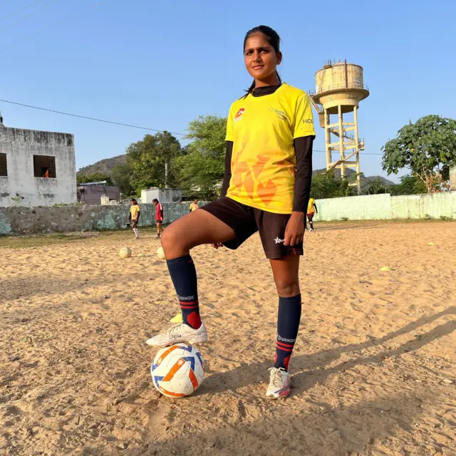 पीली टी-शर्ट और काली शॉर्ट्स पहने मुन्ना प्रैक्टिस ग्राउंड में खड़ी है और दायाँ पैर फुटबॉल पर टिकाया हुआ है.