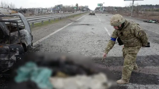 Here a Ukrainian soldier gestures towards sets of charred human remains by a burned out vehicle