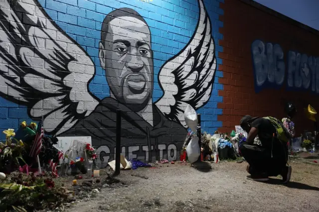 A man kneels in front of a memorial and mural for George Floyd