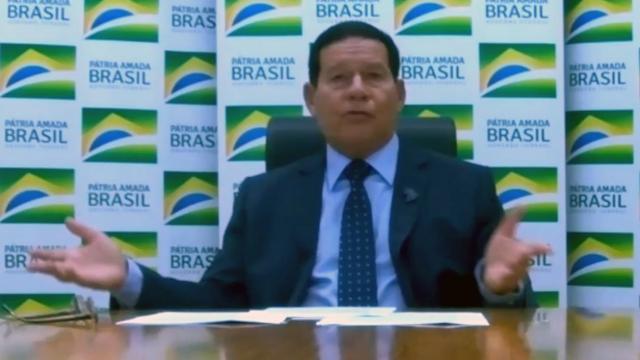Em transmissão online de vídeo, Mourão aparece sentado diante de mesa, gesticulando com braços abertos enquanto dá entrevista, com painel escrito 'Pátria amada Brasil' atrás