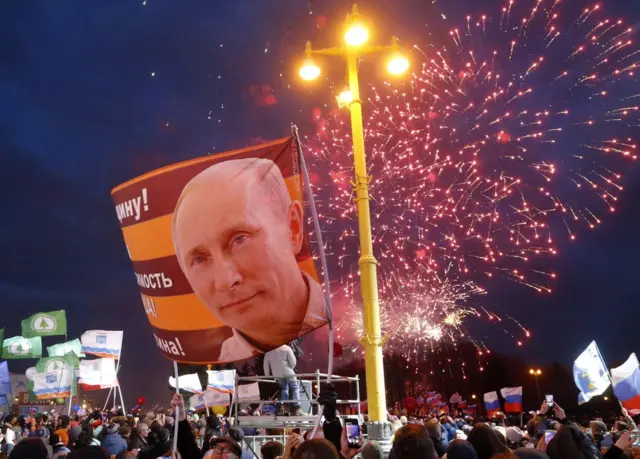 Люди держат нациоональные флаги и портреты с изображением Владимира Путина