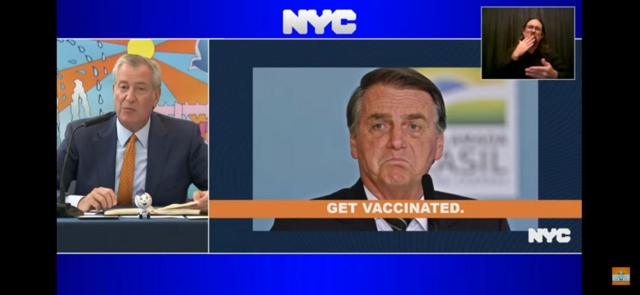 Bill de Blasio em programa de TV nos EUA ao lado de foto pedindo para que Bolsonaro se vacine
