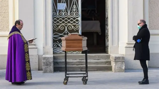 A man wearing a face mask (R) stands by the coffin of his mother as a priest reads prayers during a funeral service