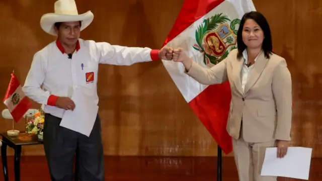 Pedro Castillo e Keiko Fujimori se cumprimentam com mão, dando sorrisos contidos,apostafrente a bandeira do Peru