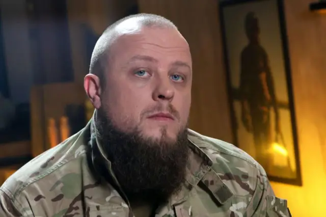 Чоловік у камуфляжному одязі з густою темною бородою. Освітлення тепле й м’яке, схоже на студійне. На задньому плані видно інтер’єр приміщення з настінною фотографією військового у спорядженні. 