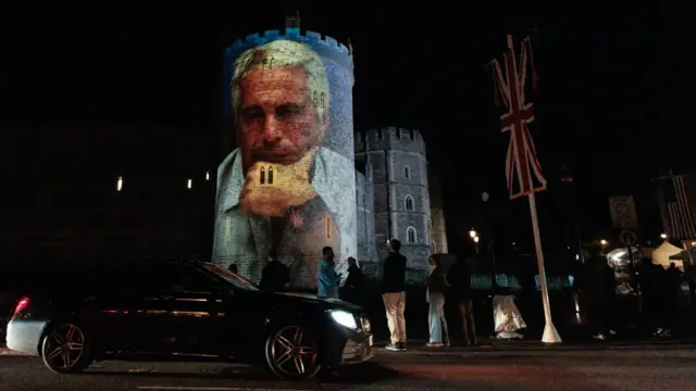 تصویری از جفری اپستین که دستش را زیر چانه گذاشته، روی برجی در قلعه ویندزور، نزدیک لندن، انداخته شده. پرچم بریتانیا از میله‌ای در همان نزدیکی آویزان است. گروهی از مردم در حال تماشای تصویر هستند، ۱۶ سپتامبر ۲۰۲۵