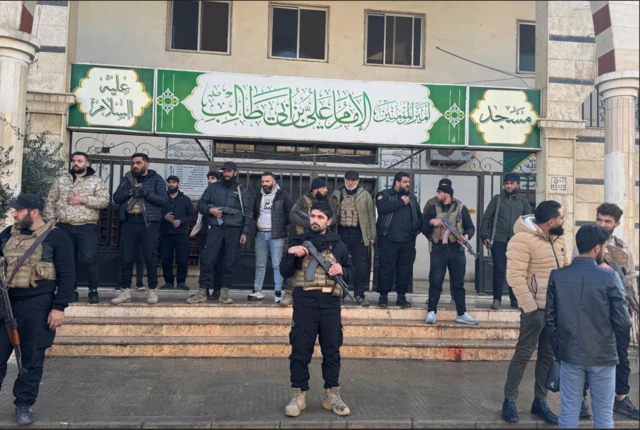 محتوى صادم: مسلحون يقفون خارج مسجد الإمام علي بن أبي طالب بجوار بقعة دماء على الدرج عقب انفجار في حي وادي الذهب بمدينة حمص في 26 ديسمبر/كانون الأول 2025.