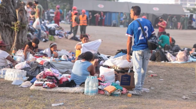 As pessoas se sentam no chão, cercadas por seus pertences, com uma tenda de emergência ao fundo.