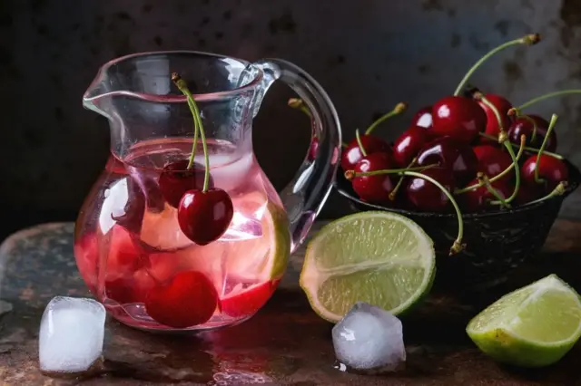 Limonade maison aux cerises et au citron vert, servie dans une carafe en verre avec des glaçons, des cerises fraîches et des tranches de citron vert sur un fond rouillé en fer foncé. 