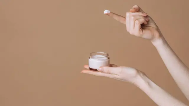 Uma mão segura um pote de creme, enquanto o conteúdo é retirado com o dedo de outra mão.
