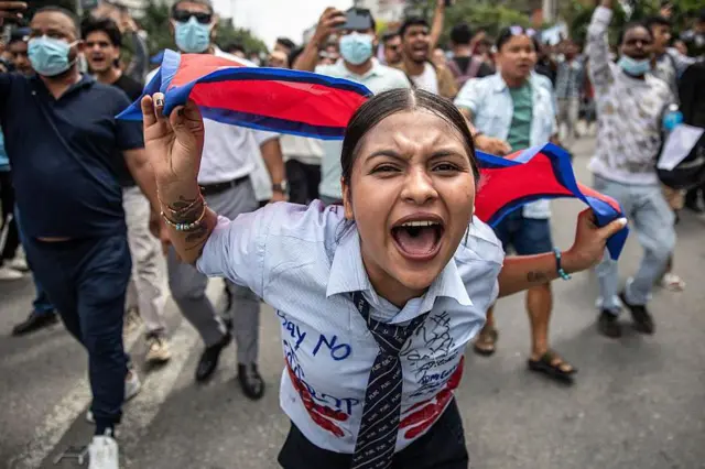 Mwandamanaji wa kike aliyeshika bendera ya Nepal akipiga kelele wakati wa maandamano nje ya Bunge huko Kathmandu tarehe 8 Septemba 2025, akilaani madai ya ufisadi wa serikali na vikwazo kwenye mitandao ya kijamii. Ameshikilia bendera nyuma ya kichwa chake na amevaa shati la sare ya shule iliyoandikwa kwa wino juu yake.