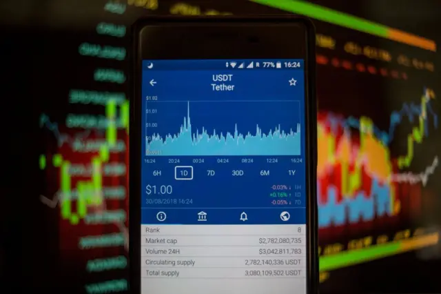 در این تصویرِ ایلوستریشن که در بانکوک تایلند و در تاریخ ۳۰ اوت ۲۰۱۸ ثبت شده، یک تلفن هوشمند در کادر دیده می‌شود. روی صفحه‌نمایش گوشی، ارزش بازار رمزارز تتر (USDT) از طریق یک اپلیکیشن معاملاتی به نام کریپتو نمایش داده شده است. اعداد و نمودارهای مالی روی صفحه، تغییرات قیمتی و وضعیت بازار را نشان می‌دهند. تمرکز تصویر بر صفحه گوشی است و پس‌زمینه حالت نمادین دارد تا فضای دیجیتال و بازار رمزارزها را القا کند.