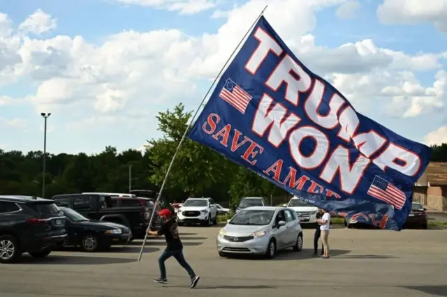 Homem caminha em estacionamento com grande bandeira que diz: Trump venceu